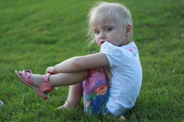 Küçük kız çimenlerde oturur ve sandalet giyer.