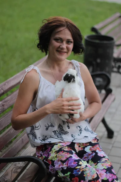 woman in a white blouse with butterflies and a colorful skirt sits on a bench with a white rabbit in her arms