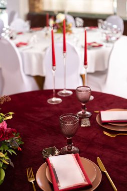 Ziyafet masasına koyun: menü kartı tabakta kırmızı bir peçete, tabağın kenarlarında yaldızlı bir çatal ve bıçak, bir bardak kahverengi cam, çiçek aranjmanı, kırmızı mumlar