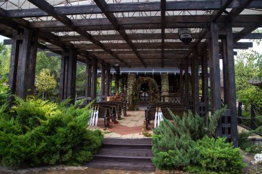 açık ahşap çadır, yeşil dallardan ve kırmızı çiçeklerden oluşan düğün kemeri, dekoratif tekerleklerdeki ahşap banklar, beyaz kamış şemsiyeler