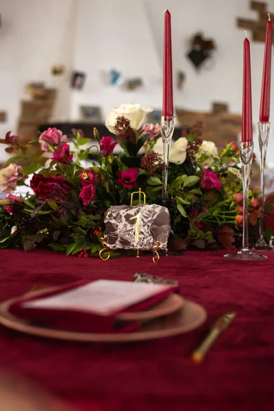 Place at the banquet table: the menu card lies on a red napkin on plates, a gilt fork and knife on the sides of the plate, a glass of brown glass, flower arrangement, red candles