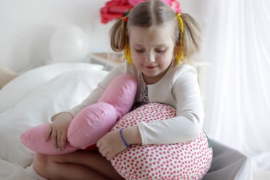 Bir kız beyaz ve pembe balonlarla ve çilek yastıkla yatakta oynuyor.