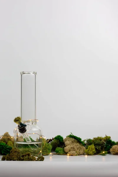 Glazen waterpijp gevuld met onkruid omgeven door wiet toppen, geassorteerde moss en string lichten tegen Wit — Stockfoto