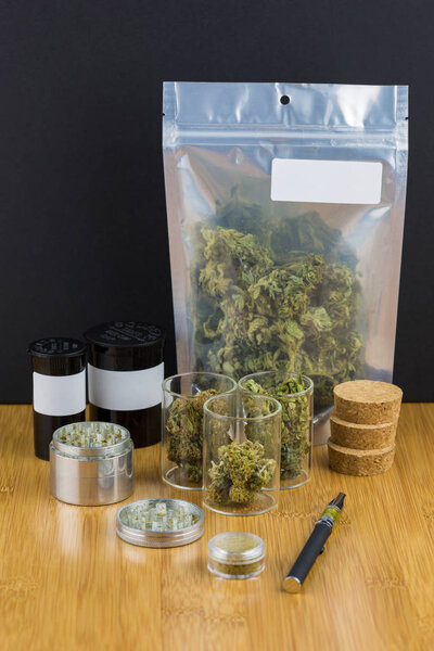 assorted medical marijuana in containers and plastic bags with metal grinder and vape pen on wood surface against black backdrop