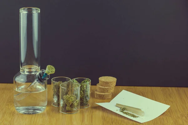 Marihuana medicinal en recipientes de vidrio junto a la pipa de vidrio con papeles de laminado en forma de cono y junta — Foto de Stock