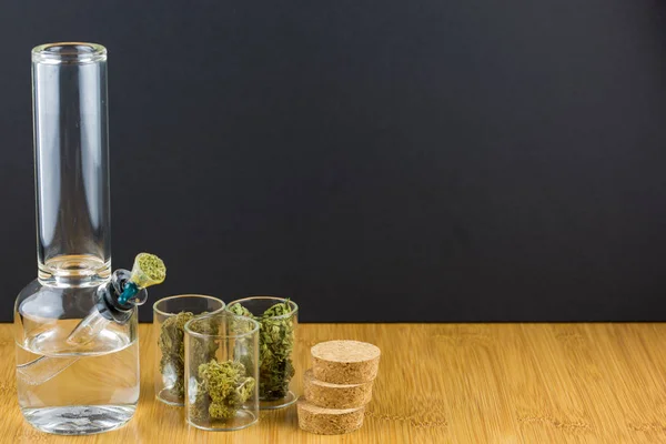 Verschiedenes medizinisches Marihuana in Glascontainern neben Glasbong auf Holzoberfläche mit schwarzem Hintergrund — Stockfoto