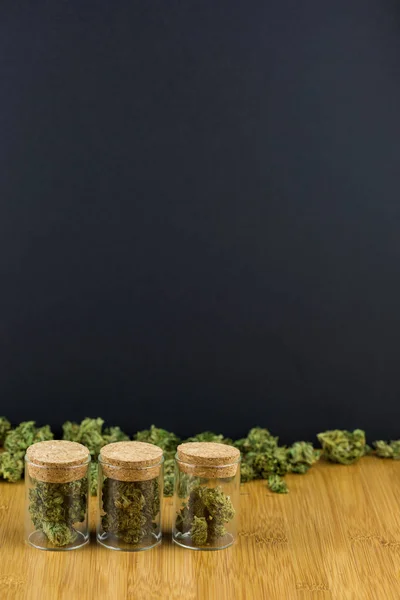 Pots en verre remplis de marijuana dans une rangée avec bourgeon lâche sur la surface du bois avec fond noir — Photo