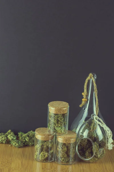 Marihuana in verschiedenen Glascontainern mit loser Knospe auf Holzoberfläche vor schwarzem Hintergrund — Stockfoto