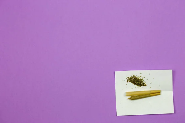 Cone shaped rolling paper with loose marijuana and joint on purple surface