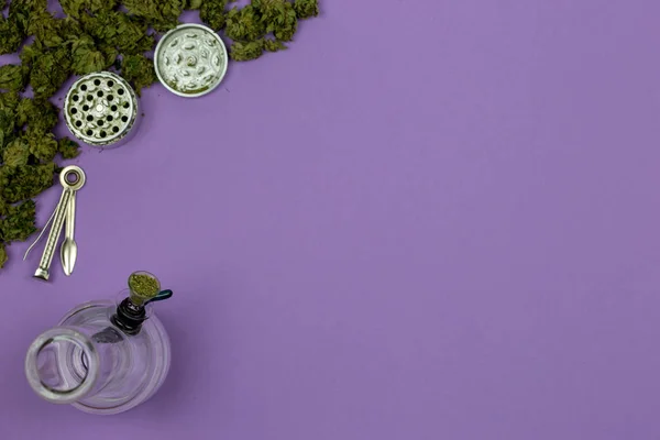 Marijuana en vrac avec bong en verre, broyeur de métal et outil de nettoyage sur la surface violette — Photo