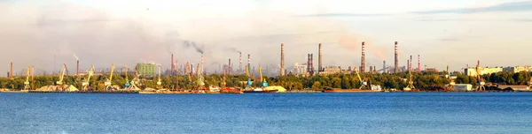 Boruları olan sanayi kompleksi, panoramik şehir manzarası. Bir nehir ve büyük metal fabrikaları binaları. Atmosferde duman salınımı.