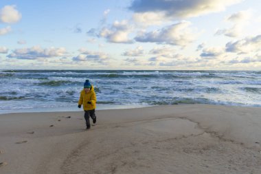 Parlak sarı ceketli bir çocuk deniz kıyısında güneşli bir sonbahar günü koşar.