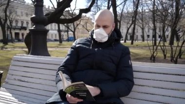 Enfeksiyon kapmış kel kafalı genç beyaz bir adam parkta solunum cihazında oturmuş kitap okuyor. 