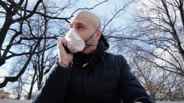 Enfeksiyon kapmış kel kafalı genç beyaz bir adam parkta solunum cihazında oturmuş telefonda hastalığını tartışıyor.