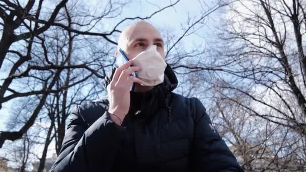 感染症の頭を持つ若い白人男性が新鮮な空気中の公園のマスクに座って電話で彼の病気を議論しています — ストック動画