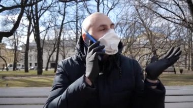 Enfeksiyon kapmış kel kafalı genç beyaz bir adam bir solunum cihazında oturuyor ve temiz havada bir parkta tıbbi eldivenler takıyor ve telefonda bir şey konuşuyor.