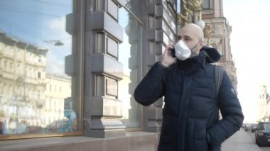 Enfeksiyon kapmış kel kafalı genç beyaz bir adam sokakta tek başına solunum cihazıyla yürüyor ve telefonla konuşuyor.