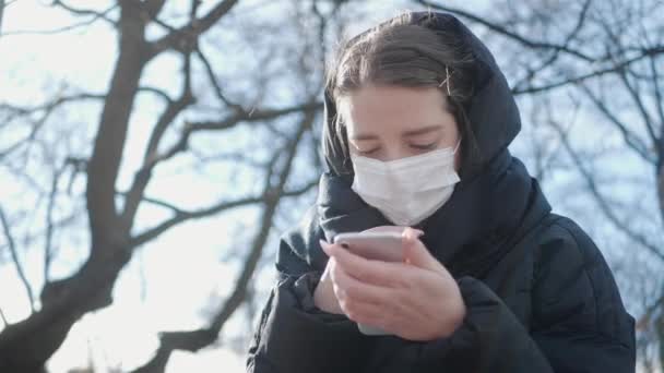 一名身穿冬季夹克 头戴医用口罩的少女坐在公园里冲破隔离带 一边在手机上留言 图库视频片段
