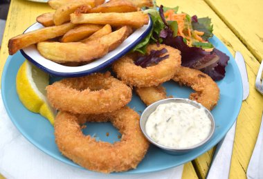 Kızarmış kalamar, salata ve patates cipsi mavi seramik tabakta ve sarı masa arkasında..