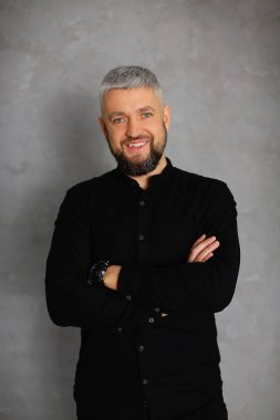 Siyah giyinmiş, gri arka planda saati olan yaratıcı sakallı bir beyefendinin yakın plan portresi.