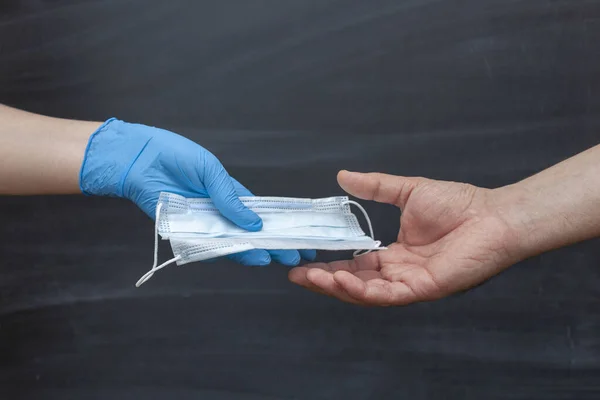 Glove hand gives a medical face mask to hand another person to protect against viruses during an epidemic. Chalk Board in the background