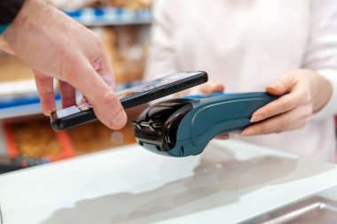 Bir kadının elleri ödeme terminali tarafından tutulur ve bir erkek akıllı telefon ile satın almak için ödeme yapar. Yan görüş. NFC, iş ve bankacılık işlemleri kavramı.