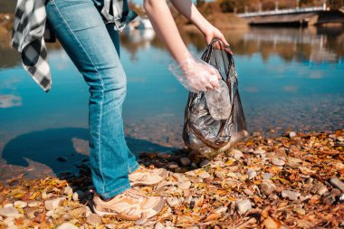 Çevre Koruma ve Toprak Günü. Bir kadın gönüllü, göl veya nehir kıyısındaki bir çöp torbasına plastik bir şişe koyar.