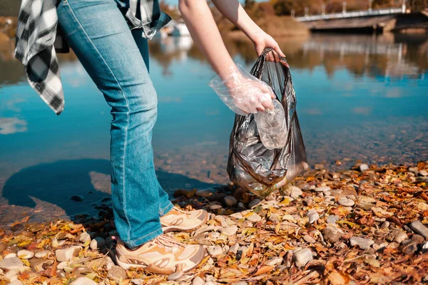 Environmental protection and earth day. A female volunteer puts a plastic bottle in a garbage bag on the shore of a lake or river