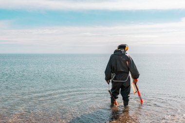 A man standing knee-deep in the water looking for precious metals with a metal detector. Sea and sky on the background. clipart