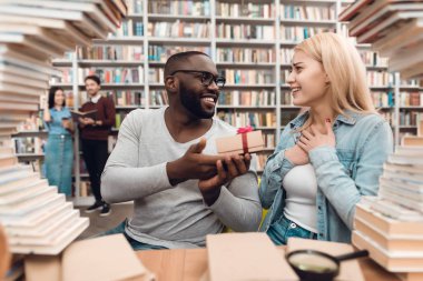 Etnik Afrikalı-Amerikalı adam ve Kütüphane masada oturan ve hediye veren beyaz kız