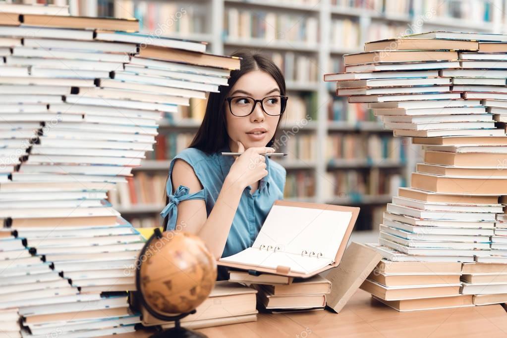 Ethnic asian girl in glasses sitting at table surrounded by books in library and writing in notebook