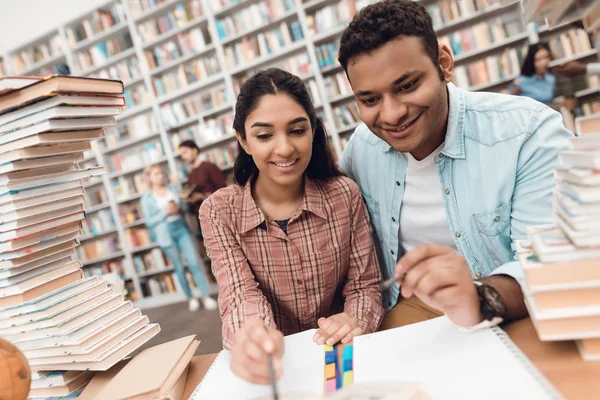 Ethnic indian mixed race girl and guy sitting at table surrounded by books in library and taking notes