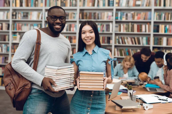 Group of ethnic multicultural students sitting at table in library, black guy and asial girl with books