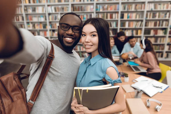 Group of ethnic multicultural students sitting at table in library, black guy and asian girl taking selfie on smartphone
