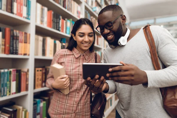 Ethnic indian mixed race girl and black guy surrounded by books in library and tablet