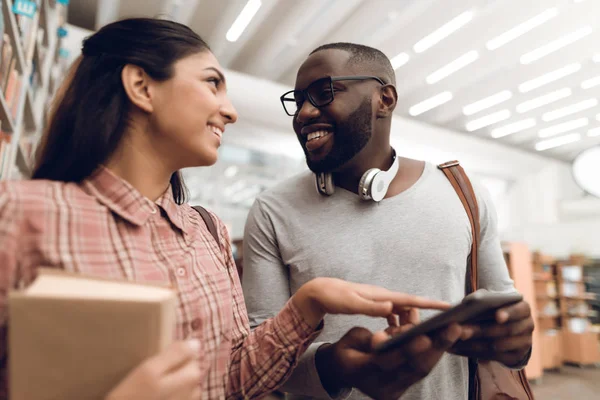 Ethnic indian mixed race girl and black guy surrounded by books in library and tablet