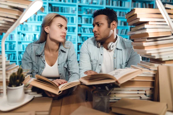 Ethnic indian mixed race guy and white girl sitting at table surrounded by books in library and reading books