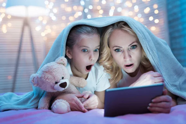 Grandmother and granddaughter watching movie on tablet under blanket on bed at home