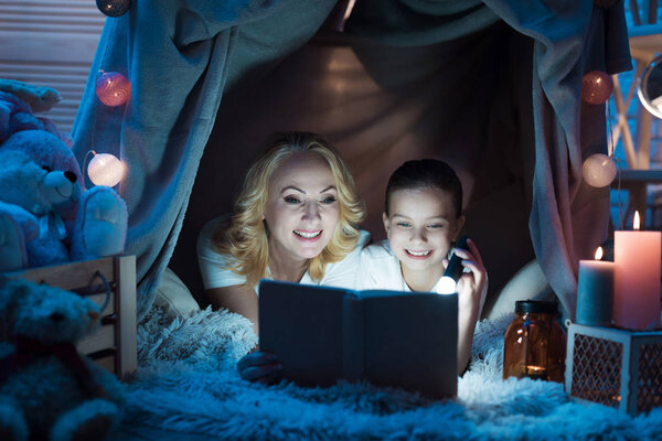 Grandmother and granddaughter reading book in blanket house with flashlight at home