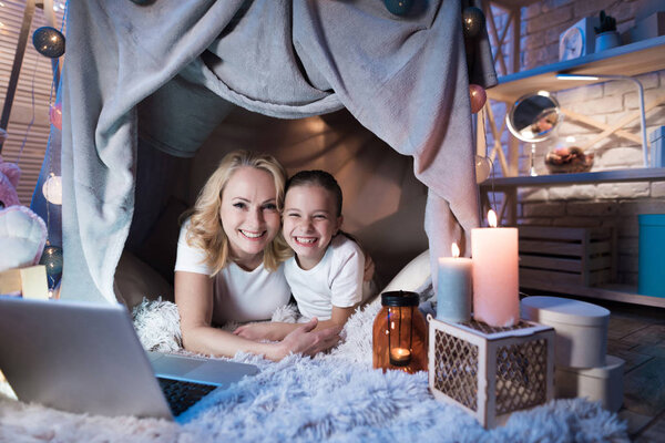Grandmother and granddaughter lying with laptop in cozy blanket house at home