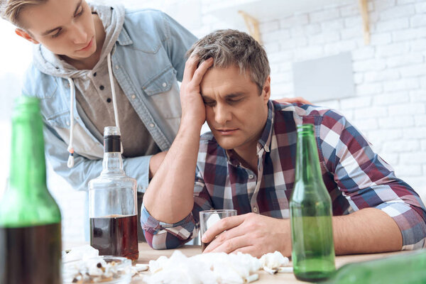 father sitting at table with glass and drinking while son trying stop him