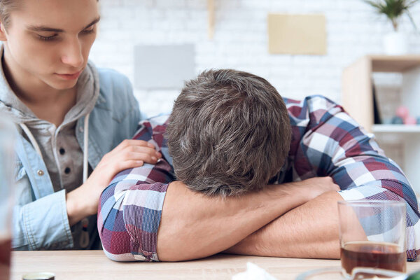 Drunk father lying on table with glass and bottles while son trying stop him