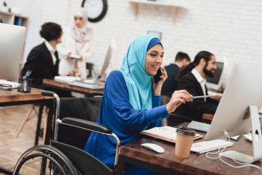 Arap kadın Türban masaüstü bilgisayarda çalışarak ve smartphone üzerinde konuşurken tekerlekli sandalyede devre dışı
