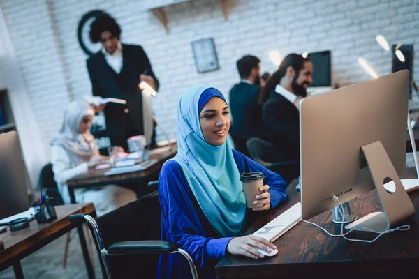 Disabled arab woman in hijab in wheelchair working on desktop computer and drinking coffee