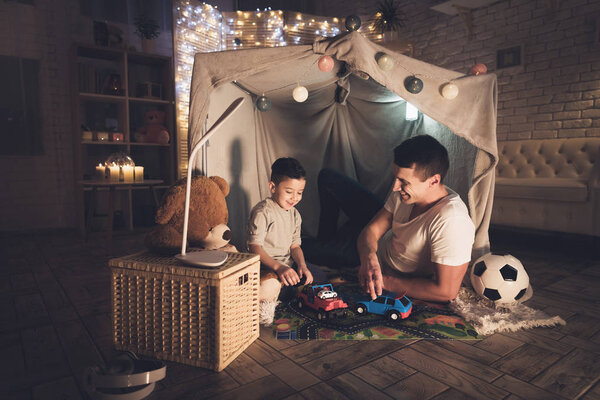 Father and son playing with toy cars on carpet road in blanket fort 