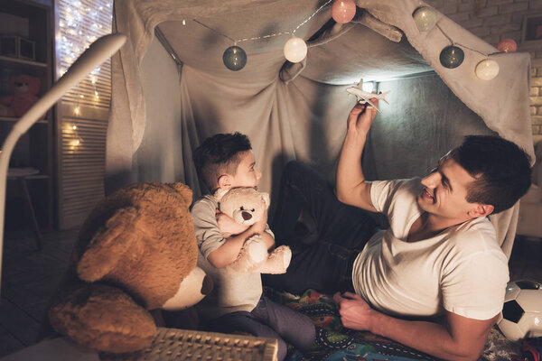 Father and son playing with toy airplane and cars in blanket fort 