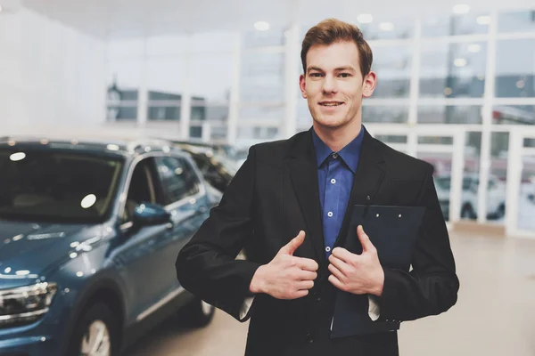 Car salesman posing with clipboard near new auto at car dealership