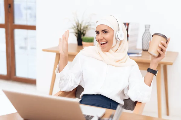 Arab woman in hijab sitting near laptop and listening music on headphones