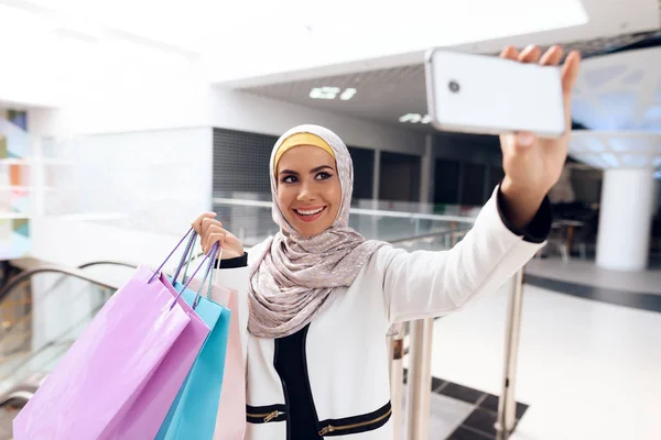 Arab woman walking in shopping mall with shopping bags and taking selfie on smartphone