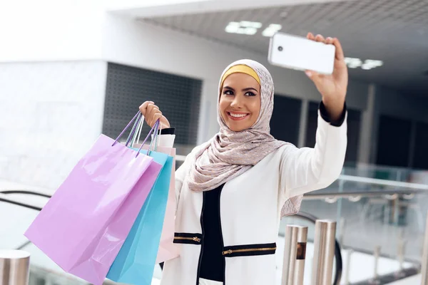 Arab woman walking in shopping mall with shopping bags and taking selfie on smartphone